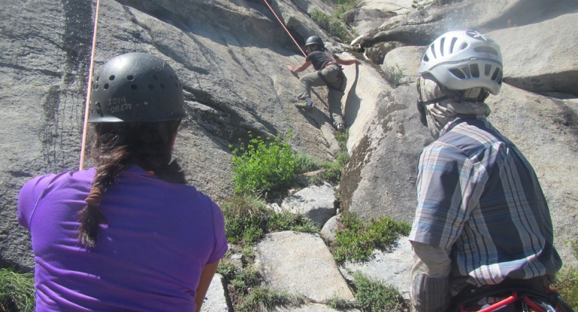 two people wearing helmets look on as another person rock climbs on an outward bound course for bipoc